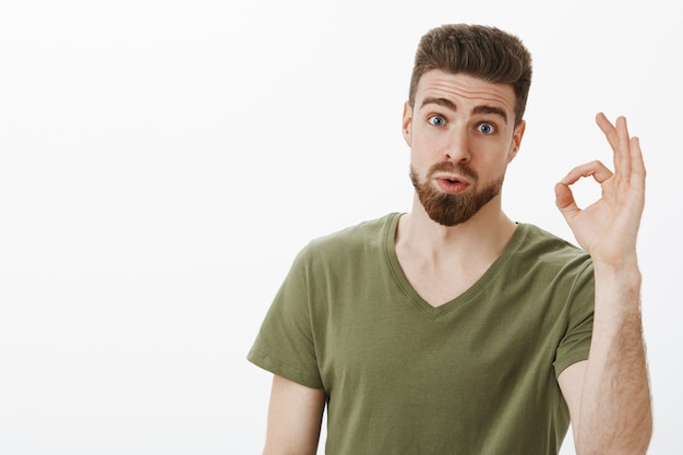 Portrait of impressed guy checking out awesome idea of friend saying wow and not bad showing okay gesture raising eyebrows as being amazed with nice plan posing astonished over white wall