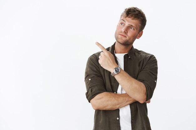 Portrait of impressed handsome male customer doing shopping tlting head curiously as pointing and looking left with interest as seeing cool thing, posing over gray wall