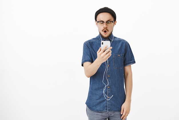 Portrait of impressed and stunned amazed good-looking adult guy with beard in glasses and beanie folding lips in omg or wow staring at smartphone screen impressed and surprised wearing earphones