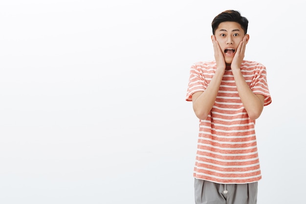 Portrait of impressed and surprised excited young asian male student screaming from amazement and joy pressing hands to cheeks and staring thrilled and astonished on right side of copy space