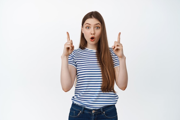 Portrait of imrpessed girl with WOW face pointing fingers up stare in disbelief at camera amazed by sale banner standing over white background