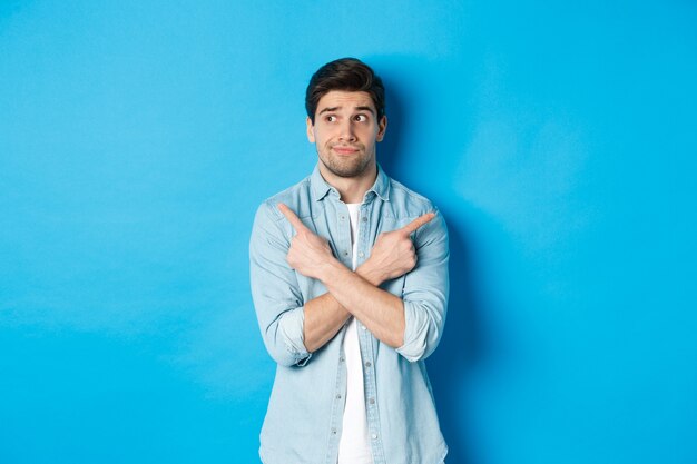 Portrait of indecisive adult man pointing fingers sideways but looking left, making choice between two products, standing against blue background.