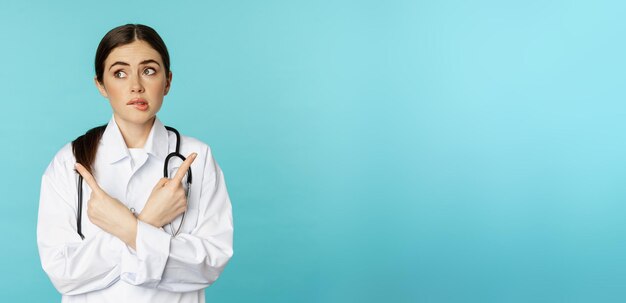 決断力のない混乱した女性医師の肖像画 薬局の労働者が横を指し,唇をみ,青い背景の上に立っていることを知らない.