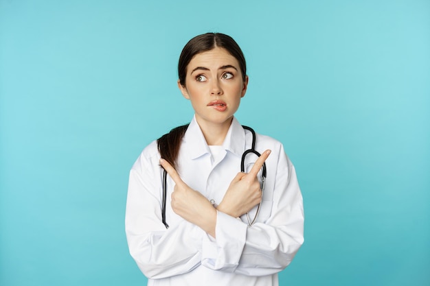 Portrait of indecisive, confused female doctor, pharmacy worker pointing sideways and biting lip clueless, dont know, standing over blue background