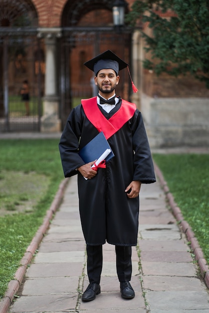 Foto gratuita ritratto di laureato indiano in abito di laurea nel campus universitario.