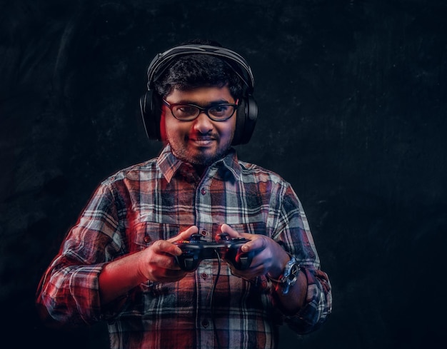 Portrait of a Indian guy in wireless headphones holding joystick and plays video games on console standing in the studio