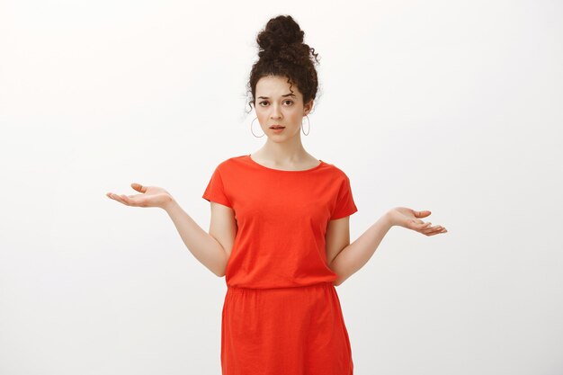 Portrait of indifferent careless Caucasian curly-haired woman in red dress, shrugging with spread hands