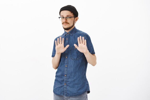 Portrait of indifferent and unimpressed handsome stylish bearded guy in glasses and black beanie raising palms in rejections and decline gesture being uninterested in strange offer