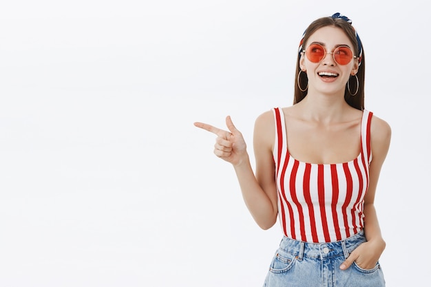 Portrait of interested creative and stylish hot woman in striped top and red sunglasses holding hand in pocket casually pointing and gazing left with enthusiastic broad smile seeing curious copy space