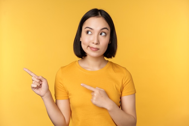 Портрет заинтригованной азиатской женщины, смотрящей и указывающей пальцем на рекламу, показывающую что-то интересное, стоящее на желтом фоне