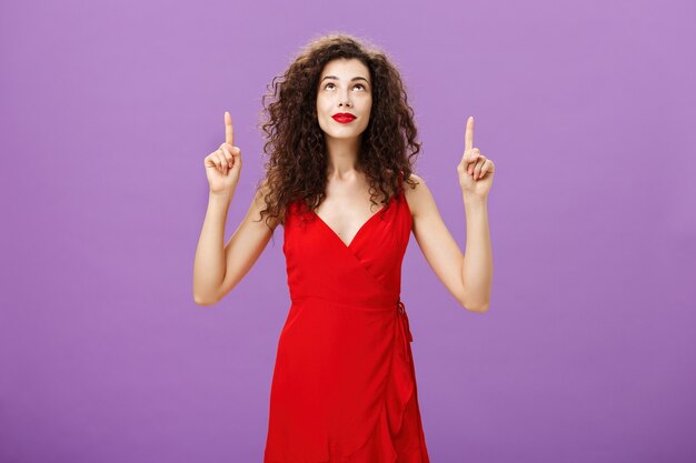 Free photo portrait of intrigued and curious charming young female with curly hairstyle in red stylish evening dress looking and pointing up smiling interested and delighted posing over purple background.