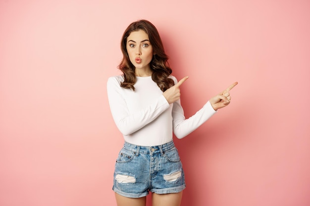 Portrait of intrigued stylish girl showing interest in advertisement, look curious, pointing at upper right corner logo, banner or discount news, pink background.