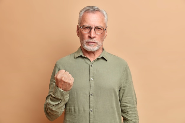 Портрет раздраженного бородатого зрелого мужчины сжимает кулак и демонстрирует гнев, предупреждает вас, одетого в строгую рубашку, позирует у бежевой стены