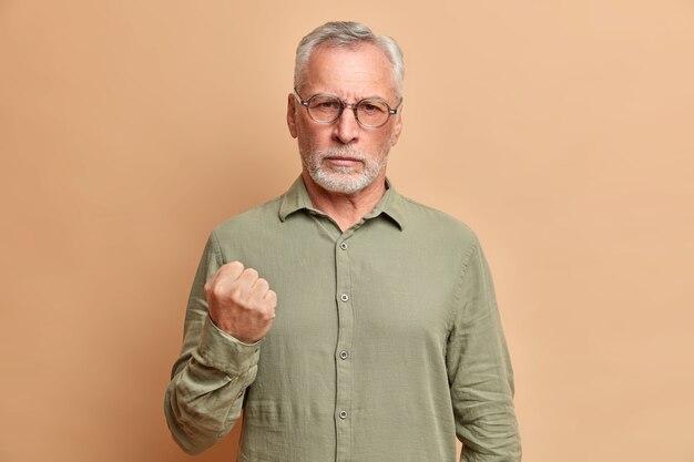 Portrait of irritated bearded mature man clenches fist and demonstrates anger warns you dressed in formal shirt poses against beige wall