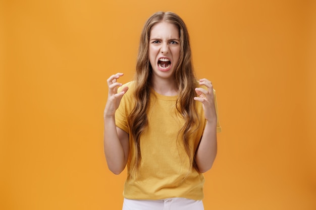 Portrait of irritated cute european female squeezing hands in fists with anger frowning and making hateful face standing annoyed and pissed over orange background with furious look. Emotions concept