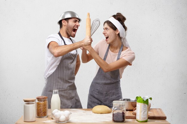 Portrait of irritated male fights with wife uses rolling pin and whisk.