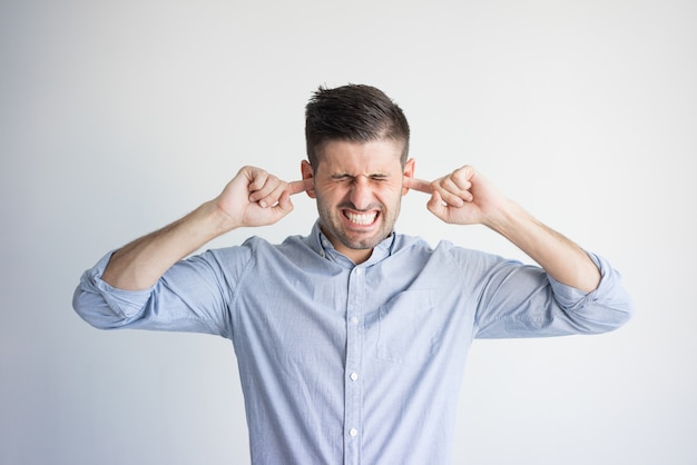 Portrait of irritated young man plugging ears with fingers.