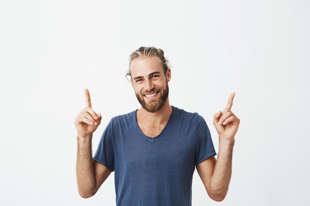 Portrait of joyful beautiful men with fashionable hairstyle and beard in blue t shirt laughing and pointing up