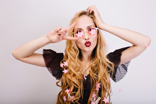 Portrait of joyful blonde with long curly hair having fun at party, making funny face, showing peace, kiss, enjoying celebration. She is wearing black dress, pink glasses. Isolated..