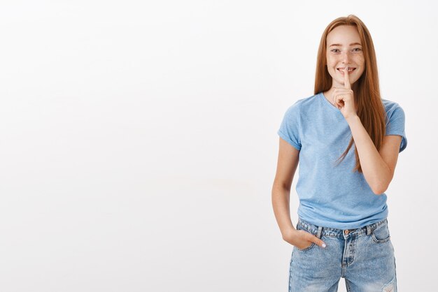 Portrait of joyful cute and carefree redhead woman in blue t-shirt and jeans showing shush gesture with index finger over mouth smiling