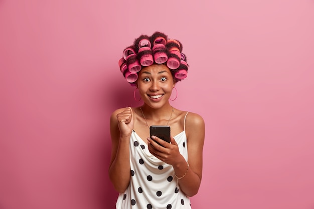 Free photo portrait of joyful dark skinned woman reads excellent news on smartphone, raises clencheed fist and smiles tootily, applies hair curlers for hairstyle. housewife uses social networks at home