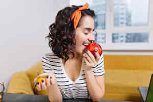 Free photo portrait joyful excited young woman with curly cut hair enjoying red apple in modern apartment. smiling, having fun, chilling at home, cosiness, relax, happiness
