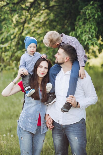 Portrait of joyful family having fun outdoors looking at camera and posing in summer park Father and mother wearing in casual clothes keeping two sons on their shoulder Concept of happy family