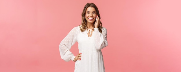 Portrait of joyful goodlooking blond woman in white dress talking on phone hold smartphone near ear and look upbeat camera smiling discuss friends date stand pink background