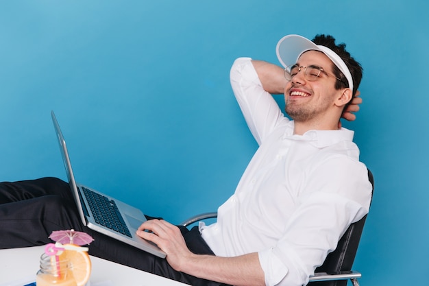 Portrait of joyful guy in glasses, white cap and shirt smiling and holding laptop.