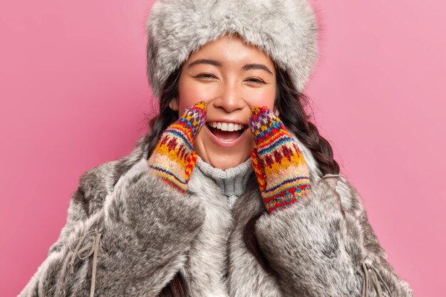 즐거운 행복한 에스키모 여자의 초상화는 겨울 코트를 착용하고 니트 장갑은 분홍색 벽 위에 고립 된 극북의 정면 생활에서 기꺼이 긍정적으로 보입니다.