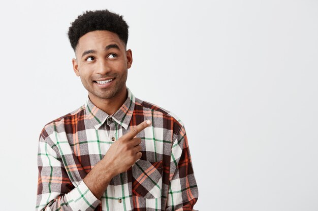 Portrait of joyful happy mature attractive black-skinned man with afro hairstyle in casual checkered shirt looking aside with raised eyebrows and smile, pointing with hand on white wall.