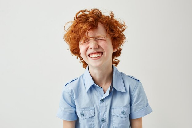 Portrait of joyful little boy with ginger hair and freckles laughing out loud with closed eyes