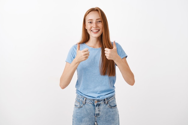 Portrait of joyful pleased good-looking redhead female with freckles showing thumbs up and smiling supportive and cheering giving positive answer