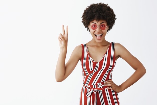 Portrait of joyful self-assured emotive and stylish african american woman in trendy sunglasses and striped overalls showing victory sign with raised hand and smiling