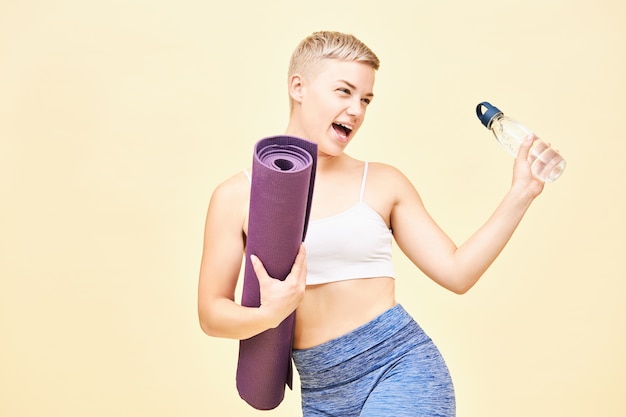 Free photo portrait of joyful sporty girl in stylish sportswear exclaiming having overjoyed facial expression, keeping mouth wide opened, feeling excited before hatha yoga class, carrying bottle and rolled mat
