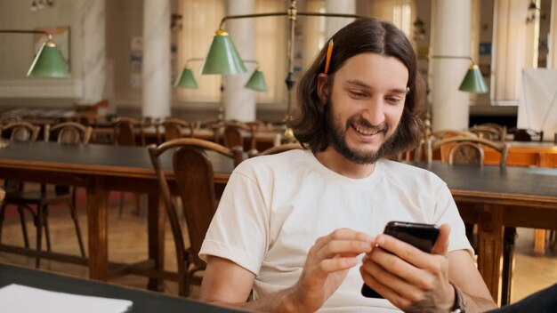 図書館でスマートフォンを楽しく使っているうれしそうな学生の肖像画大学で勉強している電話でハンサムな笑顔の男