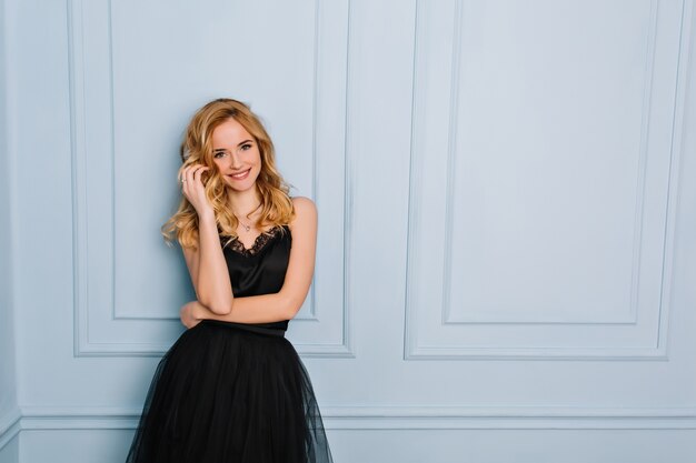 Portrait of joyful young blonde woman wearing elegant black lace dress. Blue wall. Her mouth opened, touching her face. She has long blonde wavy hair.