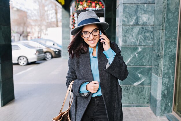 肖像画うれしそうな若い実業家の街の通りを歩いて、笑顔で電話で話します。魅力的なモデル、グレーのコート、帽子、黒のメガネ、本当の喜び。