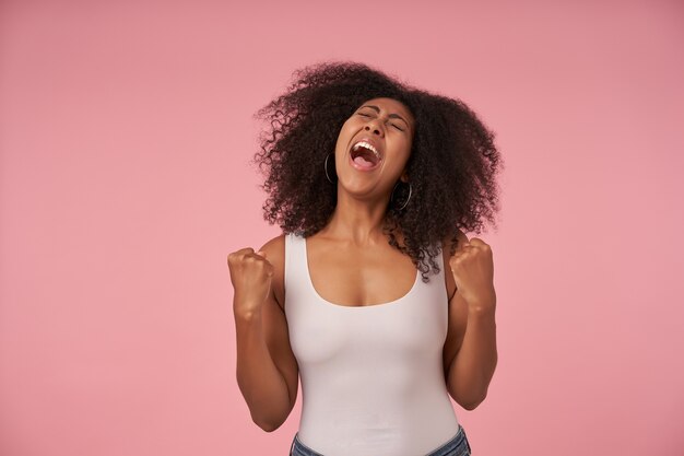 Portrait of joyful young curly female with dark skin posing on pink in white shirt and jeans, raising fists and screaming happily with closed eyes