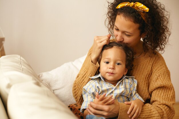 Portrait of joyful young mom in casual clothes expressing all her love and tenderness to three year old baby son, kissing him gently on forehead, spending matering leave on caring for toddler child