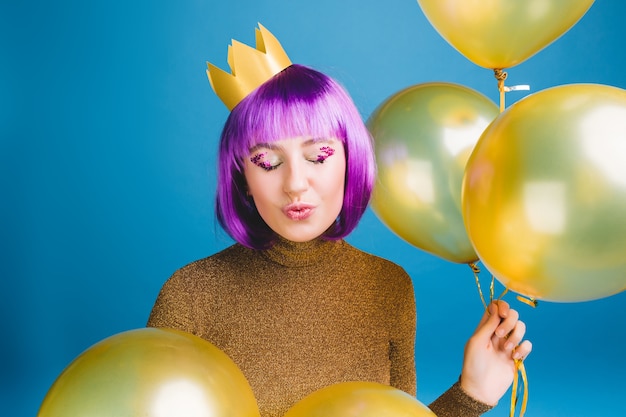 Портрет радостной молодой женщины с отрезанными фиолетовыми волосами, весело. Золотые шары, послание поцелуя с закрытыми глазами, корона на голове, роскошное платье, отличная вечеринка, праздник.