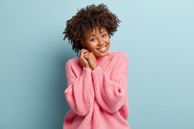 Portrait of joyous dark skinned woman has carefree facial expression, amused by friend, has gentle smile, keeps hands together near face, feels happy after date with boyfriend, has nice day.