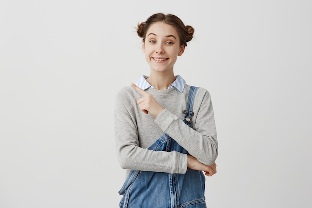 portrait of joyous young woman wearing denim pointing finger away. Positive emotions of girl gesturing on white wall proposing services. Copy space