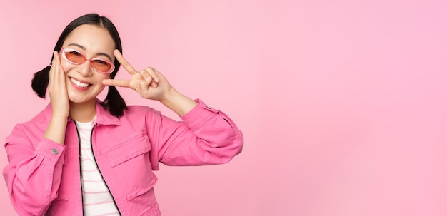 Portrait of kawaii asian girl in sunglasses showing peace vsign near eye and smiles cute at camera posing in trendy clothes against pink background