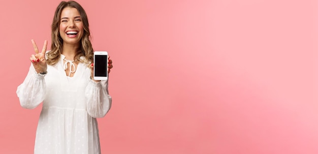 Portrait of kawaii optimistic and happy young girl in white dress show mobile phone display and peac