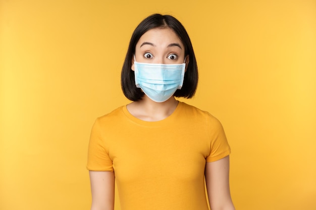 Портрет корейской девушки в медицинской маске, выглядящей удивленной, изумленной реакцией на новости, стоящие на желтом фоне
