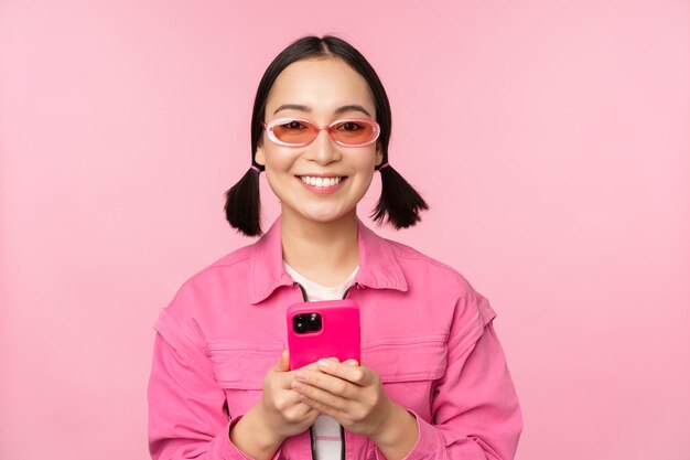 분홍색 배경 복사 공간 위에 서 있는 스마트폰 앱을 사용하여 휴대폰을 들고 있는 세련된 선글라스를 쓴 한국 소녀의 초상화