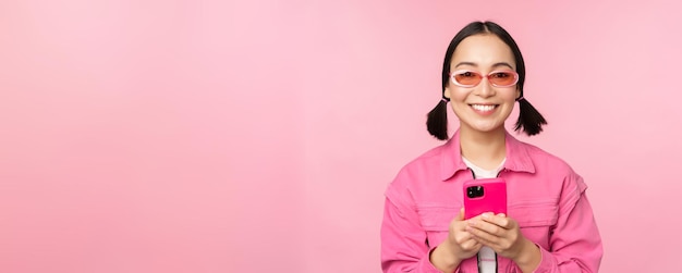 Portrait of korean girl in stylish sunglasses holding mobile phone using smartphone app standing over pink background