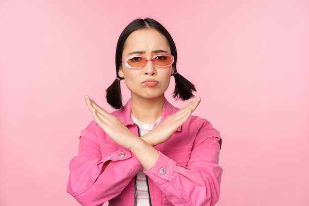 Портрет корейской девушки в стильных солнцезащитных очках, дующейся разочарованной, показывая стоп-отказ жестом креста, стоящим на розовом фоне
