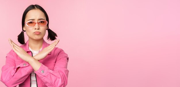ピンクの背景の上に立っている停止拒否ジェスチャークロスサインを示す失望したスタイリッシュなサングラスで韓国の女の子の肖像画